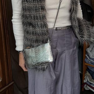 May include: A silver crossbody bag with a reptile skin texture is worn with a white turtleneck, a faux fur vest, and gray pants. A silver chain strap and a silver belt complete the outfit. A silver pendant necklace is also visible.