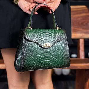 May include: A green snakeskin-textured handbag with a top handle and gold-tone hardware. The bag has a flap closure with a twist lock. The bag is held by a person wearing a black dress and a gold bracelet.
