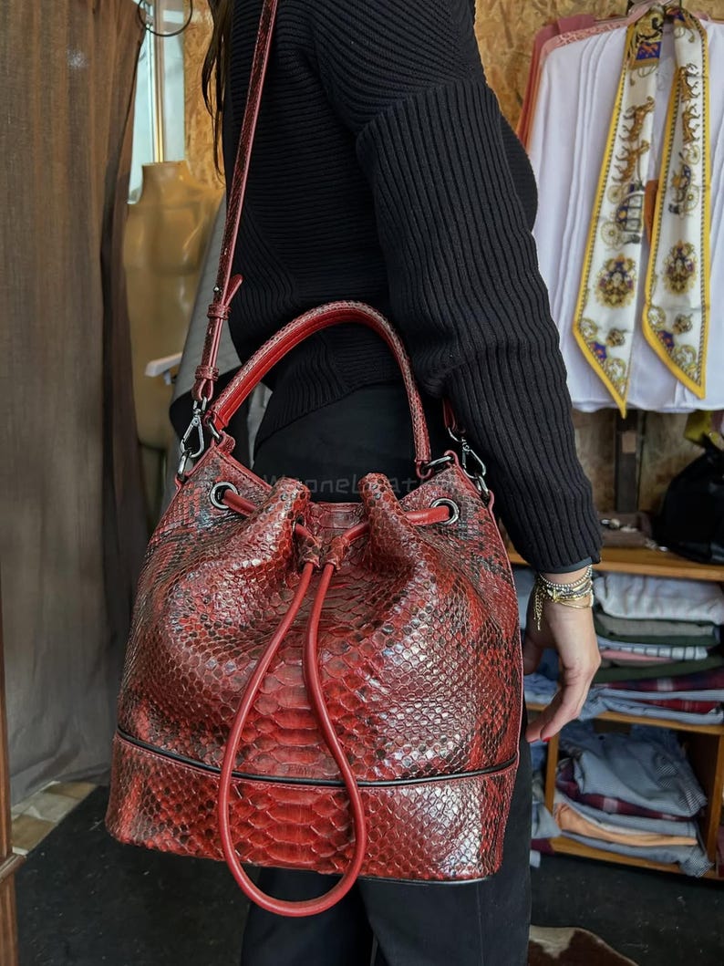 以下が含まれることがあります： 赤色のバケットバッグ、ヘビ革模様。巾着開閉、トップハンドル、ショルダーストラップ付き。バッグは質感のある素材でできており、シルバーカラーの金具が付いています。