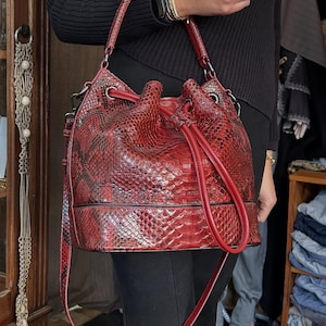 Könnte beinhalten: Roter Eimerbeutel mit Schlangenmuster, Kordelzugverschluss und langem Schulterriemen. Die Tasche hat einen strukturierten Boden und einen passenden roten Griff. Die Tasche ist tiefrot mit schwarzen Akzenten.