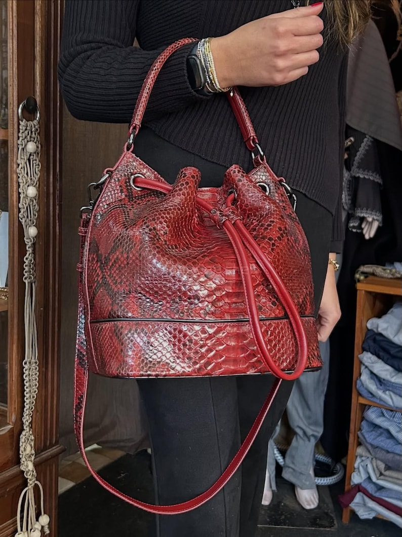 以下が含まれることがあります： 赤いヘビ柄のバケツ型バッグ。巾着開閉式で、長いショルダーストラップが付いています。バッグは構造化されたベースと、お揃いの赤いハンドルが付いています。バッグは深い赤色で、黒のアクセントがあります。