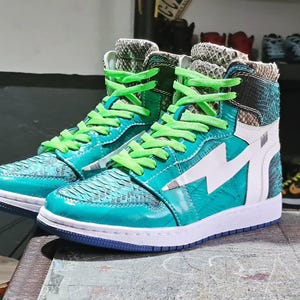 Pu&ograve; includere: Un paio di sneakers alte turchesi e bianche con un design a fulmine. Le scarpe hanno lacci verdi e una texture simile alla pelle di serpente sulla tomaia. Le suole sono bianche con la parte inferiore blu.