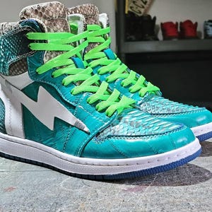 Pu&ograve; includere: Un paio di sneakers alte turchesi e bianche, con un motivo a pelle di serpente e un fulmine bianco. Le sneakers hanno lacci verde brillante e una suola bianca con bordino blu.