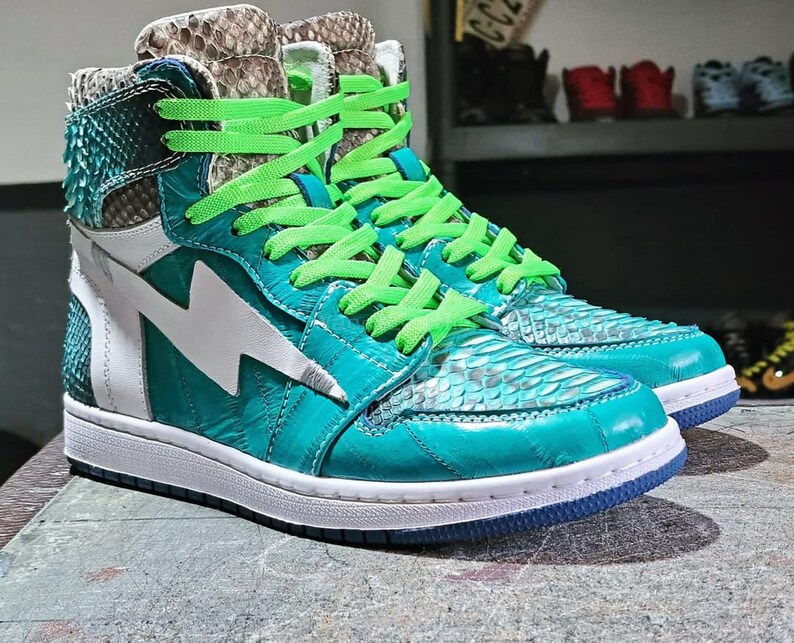 Pu&ograve; includere: Un paio di sneakers alte turchesi e bianche, con un motivo a pelle di serpente e un fulmine bianco. Le sneakers hanno lacci verde brillante e una suola bianca con bordino blu.