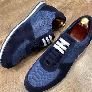 Könnte beinhalten: Ein Paar marineblaue Sneaker mit weißen Schnürsenkeln. Die Sneaker haben ein Wildleder- und strukturiertes Lederdesign. Die Sohlen sind weiß mit einem marineblauen Streifen. Die Schuhe werden auf einer Holzoberfläche präsentiert.