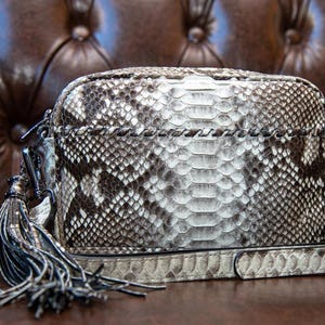 May include: A rectangular handbag with a snakeskin pattern in shades of gray and white. The bag has a long, adjustable strap and a decorative tassel. The bag is sitting on a brown surface.