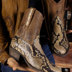 Può includere: Stivali da cowboy in pelle marrone con motivo a pelle di serpente. Gli stivali hanno la punta affilata e il tacco impilato. Il gambale è in pelle marrone più scura e il piede presenta un motivo a pelle di serpente.