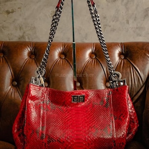 Könnte beinhalten: Eine leuchtend rote Handtasche mit Schlangenmuster. Die Tasche hat silberfarbene Kettenriemen und einen rechteckigen Verschluss. Die Tasche wird auf einem Ständer vor einem braunen Lederhintergrund präsentiert. Der Markenname "Waone Leather" ist sichtbar.