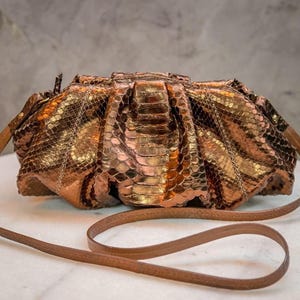 Może przedstawiać: Metaliczna kopertówka w kolorze miedzi ze skórzanym, brązowym paskiem. Torba ma teksturowany, łuskowaty wzór, a pasek jest wystarczająco długi, aby nosić go na ramieniu. Torba leży na białej powierzchni.