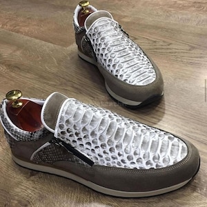 Puede incluir: Un par de zapatillas de lujo con estampado de piel de serpiente en blanco y gris. Los zapatos tienen un ribete de ante marrón, una cremallera negra y una suela marrón. Se muestran sobre una superficie de madera.