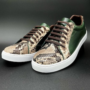 May include: A pair of leather sneakers with a snakeskin pattern on the toe and side panels. The sneakers have green leather accents, brown laces, and white soles. The shoes are displayed on a dark surface.