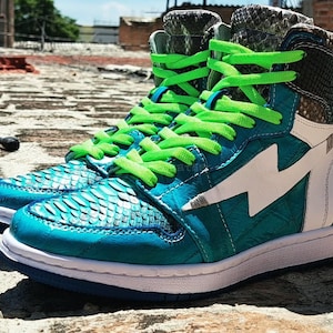 May include: A pair of high-top sneakers with teal and white accents. The sneakers feature a teal, textured upper with a white lightning bolt design and bright green laces. The heel and tongue have a snakeskin pattern.