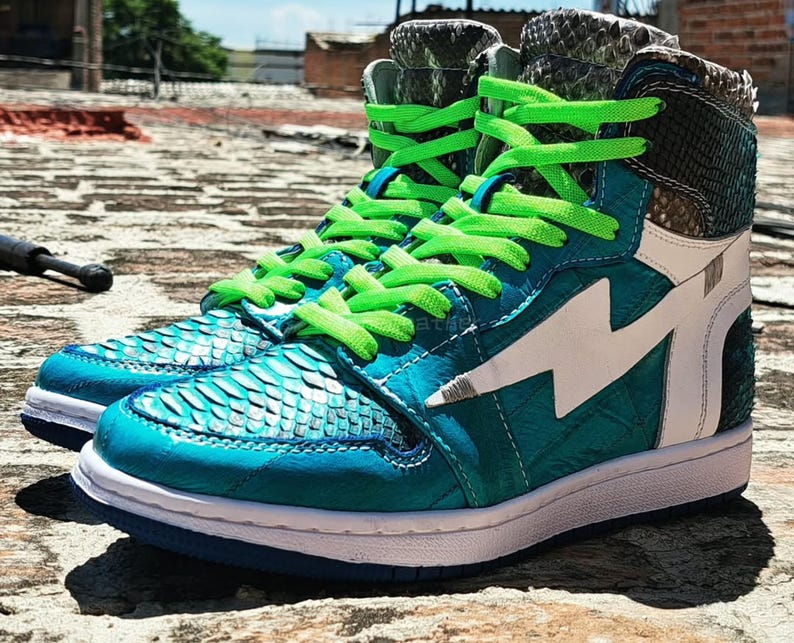 Pu&ograve; includere: Un paio di sneakers alte con dettagli turchese e bianchi. Le sneakers presentano una tomaia strutturata turchese con un design a fulmine bianco e lacci verde brillante. Il tallone e la linguetta hanno un motivo a pelle di serpente.