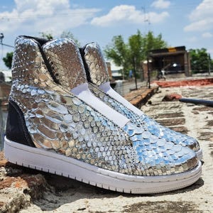 May include: High-top sneakers with a silver snakeskin pattern. The shoes have a white sole, a black heel counter, and a white elastic band across the top. The sneakers are designed with a slip-on style.