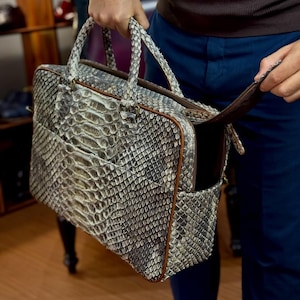 Op de afbeelding: Een rechthoekige aktetas met een slangenpatroon in grijstinten en wit. De tas heeft een bruine rits en handvatten en wordt door een persoon opengehouden. De aktetas lijkt van leer te zijn.