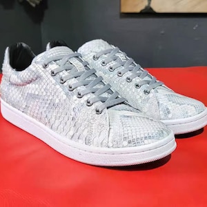 Puede incluir: Un par de zapatillas blancas y grises con un estampado de piel de serpiente. Los zapatos tienen cordones grises, suela blanca e interior negro. Las zapatillas se exhiben sobre una superficie roja.