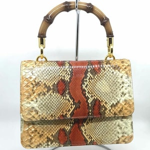May include: A rectangular handbag with a snakeskin pattern in shades of tan, brown, and red. It has a bamboo handle and gold-coloured hardware. The bag is displayed on a metal stand.
