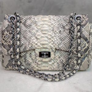 May include: A rectangular handbag with a snakeskin pattern in shades of gray and white. The bag features a silver chain strap and a silver clasp closure. The bag is sitting on a white surface.