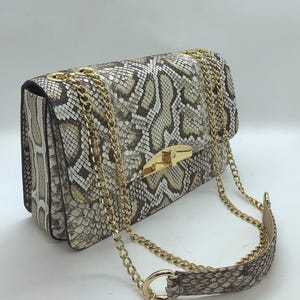 May include: A rectangular handbag with a snakeskin pattern in shades of beige, gray, and white. The bag features a gold-colored clasp and chain strap. The strap is a combination of chain and snakeskin-patterned material.