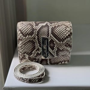 May include: A snakeskin-patterned handbag with a silver clasp and a matching strap. The bag is square and features brown and cream tones, creating a textured appearance. The strap is coiled beside the bag on a white surface.