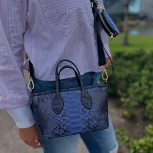 Puede incluir: Un bolso azul con estampado de piel de serpiente y una pequeña bolsa a juego. El bolso tiene dos asas pequeñas y una correa para el hombro. El bolso es de color azul oscuro con detalles en negro. La pequeña bolsa tiene cierre de cremallera y tirador dorado.