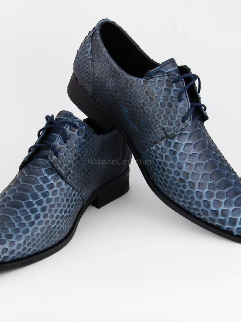 May include: A pair of blue leather dress shoes with a textured snakeskin pattern. The shoes have a pointed toe, a lace-up closure, and a low heel. The shoes are displayed on a white background.