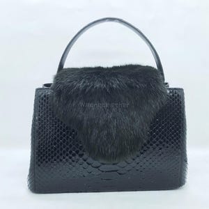 May include: A black handbag with a textured, snake-skin pattern. The bag features a short handle and a flap covered in black fur. The brand name "wagnerleather" is visible on the bag.