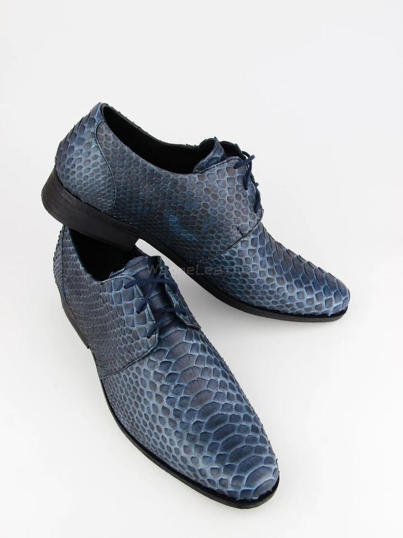 May include: A pair of blue leather dress shoes with a snakeskin pattern. The shoes have a lace-up design and a dark sole. The shoes are displayed on a white background.