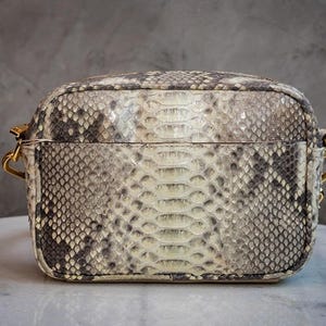 Könnte beinhalten: Eine rechteckige Handtasche mit Schlangenmuster in Grau-, Weiß- und Schwarztönen. Die Tasche hat einen Reißverschluss und goldfarbene Hardware. Die Tasche ist eine kleine Umhängetasche.