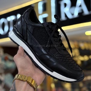 Könnte beinhalten: Schwarzer Ledersneaker mit weißer Sohle. Der Schuh hat ein strukturiertes Design an Zehen und Seitenteilen mit schwarzen Schnürsenkeln. Der Sneaker wird von einer Hand gehalten, und eine goldene Uhr ist am Handgelenk zu sehen.