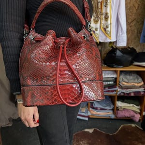 以下が含まれることがあります： 赤色のヘビ柄レザーバケットバッグ。巾着開閉式で、トップハンドルと取り外し可能なショルダーストラップが付いています。バッグの高さは約30cm、幅は約25cmです。