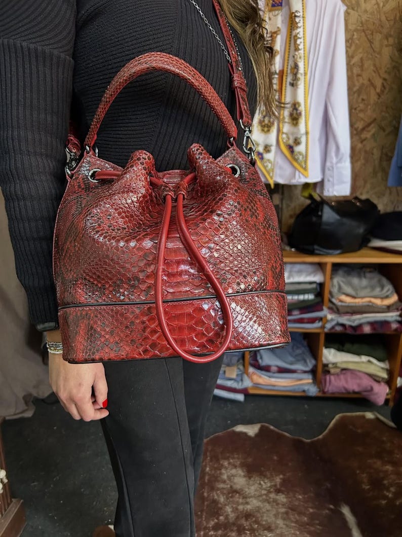 以下が含まれることがあります： 赤色のヘビ柄レザーバケットバッグ。巾着開閉式で、トップハンドルと取り外し可能なショルダーストラップが付いています。バッグの高さは約30cm、幅は約25cmです。