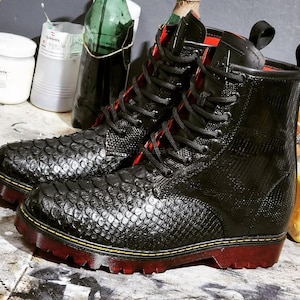 Könnte beinhalten: Ein Paar schwarze Lederstiefel mit Schlangenmuster auf der Vorder- und Seite. Die Stiefel haben rote Sohlen und Schnürsenkel, mit einer schwarzen Lasche oben. Die Stiefel stehen auf einer strukturierten Oberfläche.