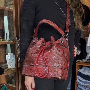 以下が含まれることがあります： 赤色のヘビ柄バケットバッグ。巾着開閉式で、お揃いのショルダーストラップが付いています。バッグにはトップハンドルとシルバーカラーの金具が付いています。バッグは深い赤色で、黒のアクセントがあります。