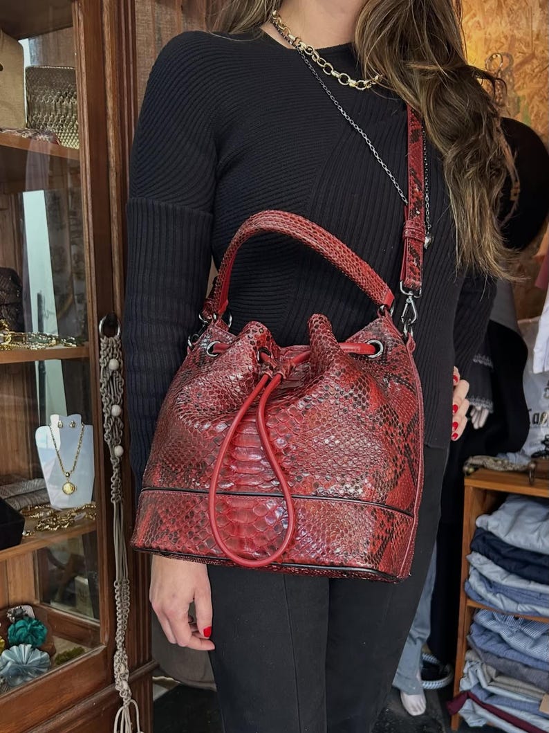 以下が含まれることがあります： 赤色のヘビ柄バケットバッグ。巾着開閉式で、お揃いのショルダーストラップが付いています。バッグにはトップハンドルとシルバーカラーの金具が付いています。バッグは深い赤色で、黒のアクセントがあります。