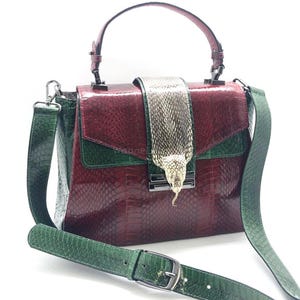 Puede incluir: Bolso de mano burdeos y verde con cierre y correa en forma de serpiente. El bolso presenta un patrón texturizado similar al de un reptil. El asa y la correa son verdes, mientras que el cuerpo principal es burdeos.