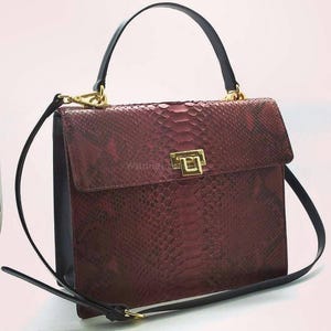 May include: A burgundy handbag with a snakeskin pattern, black trim, and a black handle. The bag has a gold-colored clasp and a detachable black shoulder strap. The bag is rectangular and has a flap closure.