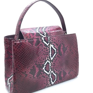 Könnte beinhalten: Eine bordeauxfarbene Handtasche mit Schlangenmuster. Die Tasche hat einen oberen Griff und eine rechteckige Form. Weiße rautenförmige Akzente sind auf der Vorderseite sichtbar. Die Tasche besteht aus einem strukturierten Material.