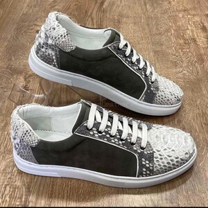 Puede incluir: Un par de zapatillas deportivas grises y blancas con un estampado de piel de serpiente en la puntera y el talón. Las zapatillas tienen un panel de ante gris oscuro y cordones y suelas blancos. Los zapatos se exhiben sobre una superficie de madera.