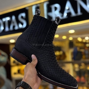 May include: Black leather ankle boot with a snakeskin texture. The boot features a square toe, stacked heel, and pull tabs. The sole is brown with white stitching. The boot is held by a hand.