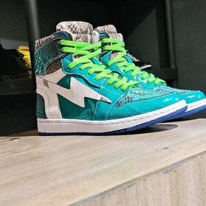 Pu&ograve; includere: Sneakers alte turchesi e bianche con un design a saetta. Le scarpe presentano lacci verdi, suola blu e un motivo a pelle di serpente sul colletto. Le sneakers sono esposte su una superficie di legno.