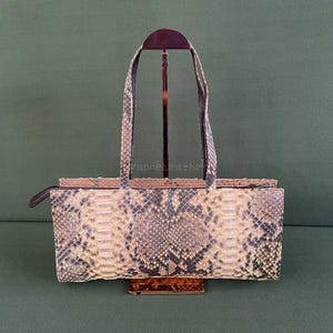 May include: A rectangular handbag with a snakeskin pattern in shades of beige, gray, and black. The bag has two long handles and a zipper closure. The bag is displayed on a brown stand against a green background.