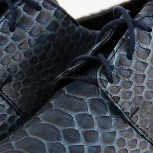 May include: Close-up of a pair of dark blue leather shoes with a scale-like texture. The shoes have matching blue laces and a classic lace-up design. The leather appears to be high-quality, with visible texture and detail.