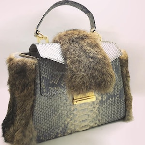 Op de afbeelding: Een luxe handtas met een slangenpatroon in grijstinten en wit, geaccentueerd met bruine bontafwerking. De tas heeft een goudkleurige sluiting en handvatbevestigingen, en een bijpassend slangenhuiddragers.