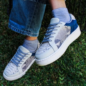 Puede incluir: Zapatillas deportivas azules y grises con un estampado de piel de serpiente en los paneles de la puntera y laterales. Las zapatillas tienen detalles de ante azul, suelas blancas y cordones grises. Las zapatillas se llevan con vaqueros azules y calcetines grises sobre hierba.