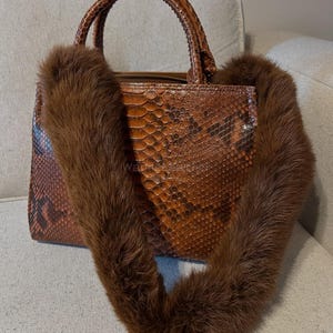 May include: A brown snakeskin-patterned handbag with a fur handle. The bag has a rectangular shape with a zipper closure and two short handles. The fur handle is a dark brown color.