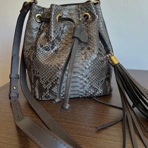 Könnte beinhalten: Graue Bucket Bag mit Schlangenmuster. Die Tasche hat einen Kordelzugverschluss mit goldfarbenen Ösen und einen langen, verstellbaren Schulterriemen. Eine Quaste hängt an der Seite. Die Tasche steht auf einer Holzoberfläche.