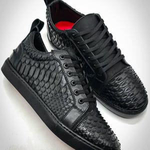 Puede incluir: Un par de zapatillas de cuero negro con un patrón texturizado en forma de escamas. Los zapatos tienen cordones negros, suelas y una lengüeta en el talón. El diseño incluye una plantilla roja.