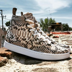 May include: High-top sneakers with a snakeskin pattern in shades of brown, tan, and white. The shoes have white laces and a white sole. The sneakers are made of a textured material and appear to be leather.