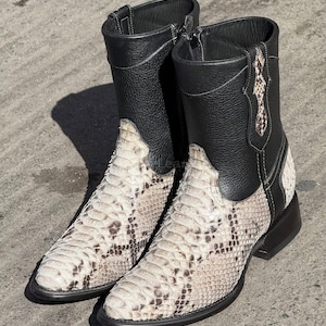 Puede incluir: Un par de botas de vaquero con un estampado de piel de serpiente en el pie y una parte superior de cuero negro. Las botas tienen cierre de cremallera y tacón apilado. El estampado de piel de serpiente es una mezcla de beige, marrón y blanco.